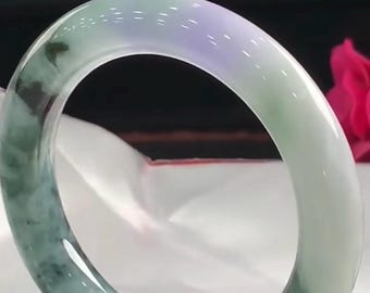 Puede incluir: Un brazalete de jade pulido y liso con una base blanca translúcida, manchas verdes moteadas prominentes y un sutil tono lavanda en la curva superior, acentuado por un pequeño cordón rojo atado en la sección inferior.