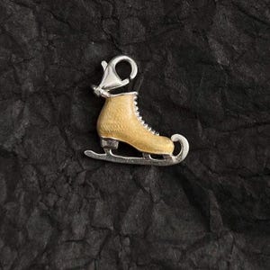 May include: A silver and gold-coloured ice skate charm. The charm features a gold-coloured skate boot with silver-coloured laces and a silver blade. The charm has a silver clasp at the top for easy attachment to a bracelet or necklace.