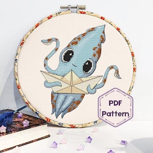 Op de afbeelding: Kruissteek hoepelkunst met een blauwe inktvis die een papieren boot vasthoudt. De inktvis heeft grote ogen en is geborduurd op crèmekleurige stof. De hoepel is versierd met een bloemenpatroon. PDF-patroonpictogram zichtbaar.