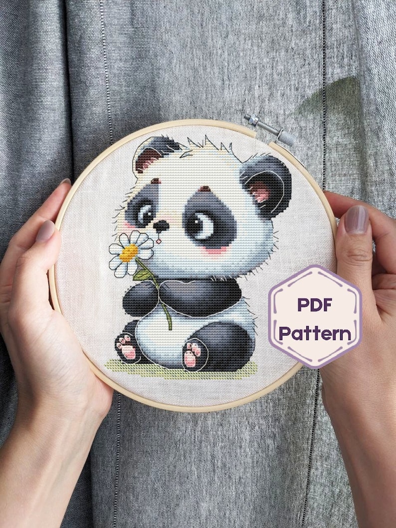 May include: Cross stitch pattern of a cute panda bear holding a daisy. The pattern is in a wooden embroidery hoop and says "PDF Pattern" in a purple hexagon.