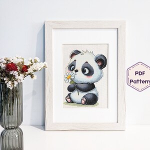 May include: A black and white panda bear cross stitch pattern with a flower in its paw. The pattern is framed in a white frame and is ready to be stitched.