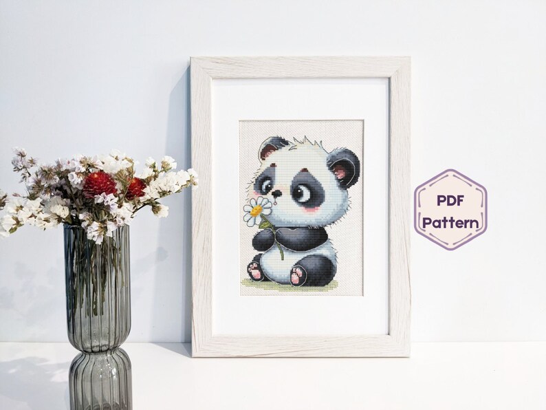 May include: A black and white panda bear cross stitch pattern with a flower in its paw. The pattern is framed in a white frame and is ready to be stitched.