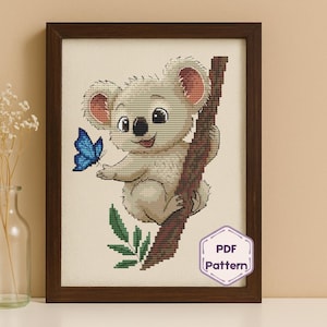 May include: A framed cross-stitch pattern featuring a cartoon koala bear clinging to a tree branch. The koala is light gray with pink accents, and a blue butterfly is nearby. The frame is dark brown, and the pattern includes the text "PDF Pattern".