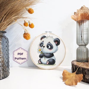 May include: A cross-stitch pattern featuring a cute cartoon panda holding a white flower. The pattern is displayed in a round wooden embroidery hoop.