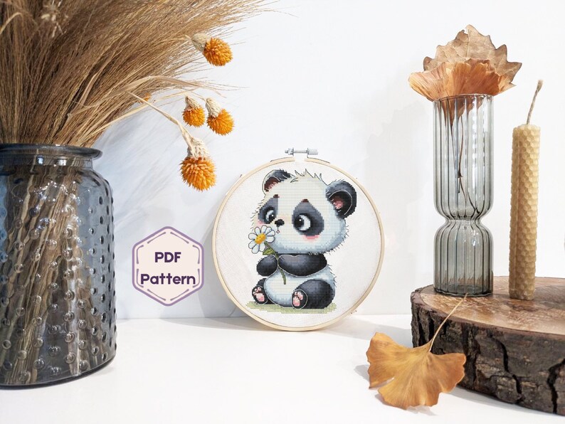 May include: A cross-stitch pattern featuring a cute cartoon panda holding a white flower. The pattern is displayed in a round wooden embroidery hoop.