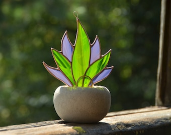 May include: A stained glass plant sculpture with green and purple leaves in a gray concrete pot. The sculpture is on a wooden surface.