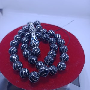 Può includere: Un rosario di perle nere con dettagli argentati e un pendente in argento. Le perle sono rotonde e hanno un motivo a strisce. Il rosario è su una superficie di velluto rosso.