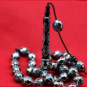 Può includere: Un mala da preghiera nero con accenti argentati e dettagli in smalto blu e bianco. Le perle sono infilate su un cordoncino nero con un fiocco nero. Il mala è su uno sfondo rosso.