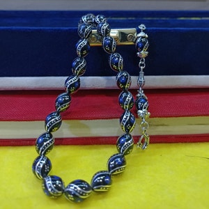 Può includere: Una collana di preghiera in perle nere con accenti blu e argento. Le perle sono di forma ovale e hanno un motivo decorativo. La collana è su uno sfondo blu e rosso.