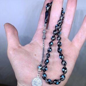 Può includere: Un rosario nero e argento con accenti blu. Le perle sono infilate su una catena d'argento con un nappa d'argento. Le perle sono di forma ovale e hanno un design argentato.