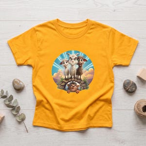 May include: A mustard yellow short-sleeve t-shirt featuring a circular graphic print of three goats standing on a stone bridge with a troll peering out from underneath. The background of the illustration shows a blue sky with clouds and sun rays.