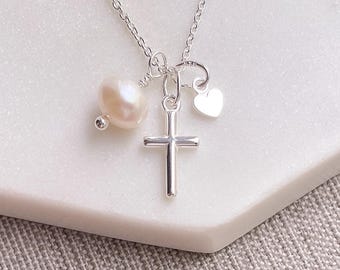 Può includere: Una collana d'argento con un ciondolo a forma di croce, un piccolo ciondolo a cuore e una perla d'acqua dolce. La collana è esposta su una superficie bianca con uno sfondo in tessuto grigio. La croce ha un design semplice e classico.