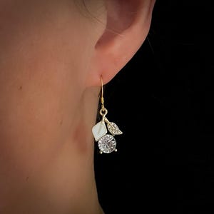 May include: Gold-tone drop earrings with a white leaf, a clear round gemstone, and a small leaf with clear crystals. The earrings have a hook closure.