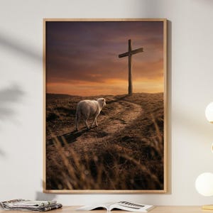 Könnte beinhalten: Ein gerahmter Kunstdruck zeigt ein Lamm, das auf einen Hügel mit einem Holzkreuz zugeht, vor einem Sonnenuntergangshimmel. Das Kunstwerk hängt über einem hellen Holzschrank mit einer goldenen Lampe und dekorativen Gegenständen.