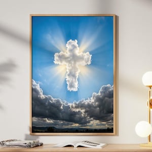 Könnte beinhalten: Ein gerahmter Kunstdruck mit einer kreuzförmigen Wolkenformation vor einem hellblauen Himmel mit Sonnenstrahlen. Das Kunstwerk befindet sich über einem hellen Holzschrank mit einer goldenen Lampe und dekorativen Objekten.
