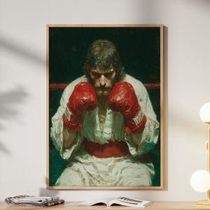 Könnte beinhalten: Ein gerahmtes Kunstwerk zeigt einen Boxer in einem weißen Gewand und roten Handschuhen vor dunklem Hintergrund. Das Kunstwerk wird über einem hellen Holzschrank mit einer goldenen Lampe und einer dekorativen Skulptur ausgestellt.