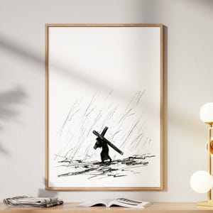 Op de afbeelding: Een zwart-wit kunstwerk in een lijst dat een figuur toont die een kruis in de regen draagt. Het kunstwerk hangt boven een licht houten kast met drie deuren. Een gouden lamp met bolvormige bollen en een decoratieve sculptuur staan op de kast.