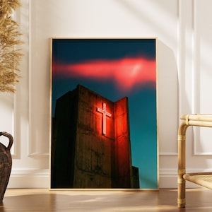 Könnte beinhalten: Ein gerahmter Kunstdruck mit einem Gebäude mit einem leuchtenden roten Kreuz. Der Himmel ist dunkelblau und rot. Das Gebäude besteht aus dunklem, strukturiertem Beton. Der Rahmen ist goldfarben.