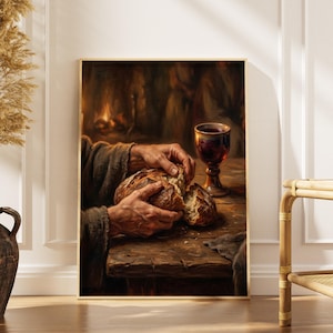May include: A framed print depicts hands breaking bread on a wooden table, with a glass of red wine. The artwork has a warm, rustic color palette, with a focus on the textures of the bread and the aged wood. The background suggests a dimly lit interior.
