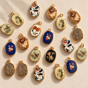 Puede incluir: Una colección de colgantes ovalados de metal dorado con varios diseños impresos, incluyendo motivos florales, pájaros, patrones de cachemira y retratos de figuras, cada uno con un pequeño lazo circular en la parte superior.