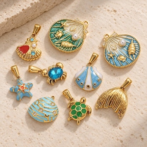 Könnte beinhalten: Eine Sammlung von goldfarbenen Metallanhängern mit nautischen Motiven, darunter zwei große Muschelanhänger mit Emaille- und Perlenakzenten, ein blauer Krebs, ein Seestern, eine Meeresschildkröte, eine Meerjungfrauenflosse und verschiedene Muschelformen.