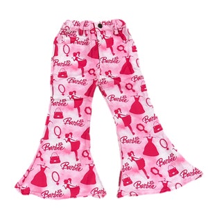 Op de afbeelding: Roze broek met wijde pijpen met een Barbie-print. De broek heeft een roze achtergrond met witte contouren van Barbie-poppen, handtassen en jurken. Het woord "Barbie" wordt herhaald in de hele print.