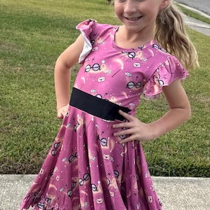 May include: A pink dress with a black waistband and ruffled hem. The dress features a repeating pattern of glasses, lightning bolts, and envelopes. The sleeves have a ruffled design. The girl is smiling and standing outside on a sidewalk.