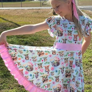May include: A light blue dress with a repeating pattern of a cartoon character and flowers. The dress has short, ruffled sleeves, a pink waistband, and a pink, ruffled hem. A pink ribbon is in the hair.