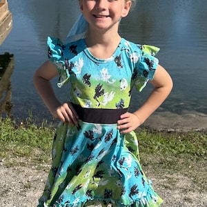 May include: A young girl wearing a blue and green dress with a black belt. The dress has a print of black and white dragons. The girl is standing in front of a body of water.