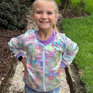 May include: A young person wearing a white iridescent jacket with a zipper closure and a colorful sequin pattern. The jacket has a purple lining. The person is wearing blue denim shorts.