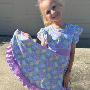 May include: A young girl wearing a purple and blue dress with a Care Bears print. The dress has a purple ruffle trim and a purple sash. The girl is smiling and holding the skirt of the dress.