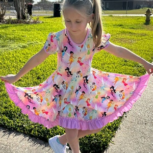 May include: A young girl wearing a pink dress with a print of fairies. The dress has a ruffled hem and short sleeves. The girl is standing on a pavement with green grass in the background.