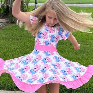 May include: A young person wearing a pink and blue dress with a cartoon print of Stitch and Angel from the Disney movie Lilo & Stitch. The dress has a pink ruffle trim and a white background.