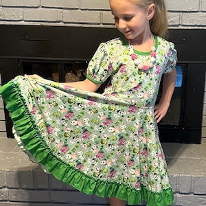 Op de afbeelding: Een lichtgroene jurk met een patroon van cartoonkatten met hoeden. De jurk heeft korte mouwen met groene bies, een groene gefranjerde zoom en een roze strik. Een kralenketting is ook zichtbaar.