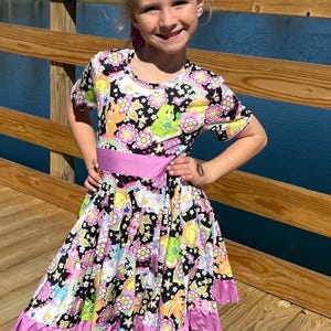 May include: A young girl wearing a black and white dress with a rainbow, stars, and cartoon bears print. The dress has a pink ruffle trim and a pink sash. The girl is wearing a pink headband and is smiling.