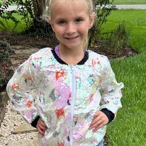 May include: A white sequined jacket with a zipper closure and a colorful print of cartoon cats. The jacket has black ribbed cuffs and a black collar.