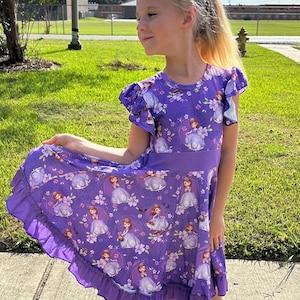 Könnte beinhalten: Ein lilafarbenes Kleid mit einem Rüschen-Saum und kurzen Ärmeln. Das Kleid hat einen Aufdruck einer Comic-Prinzessin in einem hellblauen Kleid mit einer Krone und Blumen.