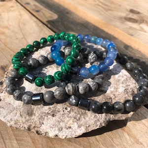 Op de afbeelding: Drie kralen armbanden gestapeld op een rots. De bovenste armband is blauw, de middelste armband is groen en de onderste armband is grijs.