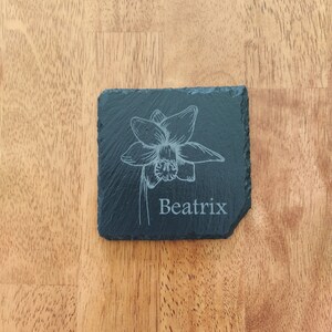 May include: Black slate coaster with a white engraved daffodil and the name Beatrix.