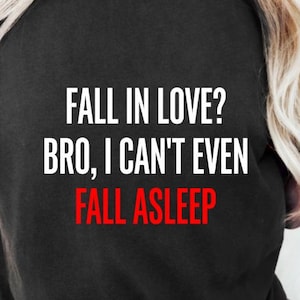 Puede incluir: Una camiseta negra de manga corta con texto impreso en blanco y rojo en la parte delantera. El texto dice: FALL IN LOVE? BRO, I CAN'T EVEN FALL ASLEEP. La prenda cuenta con un cuello redondo y un corte estándar para un estilo casual.