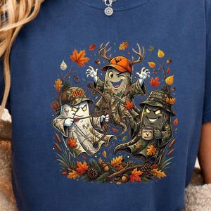 Puede incluir: Una camiseta azul marino de manga corta con un estampado gráfico de tres fantasmas de dibujos animados con ropa de caza de camuflaje y sombreros, rodeados de hojas de otoño, piñas y ramas. El fantasma central sostiene un arco.