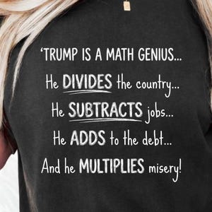 Puede incluir: Una camiseta negra con texto blanco que dice: TRUMP IS A MATH GENIUS... He DIVIDES the country... He SUBTRACTS jobs... He ADDS to the debt... And he MULTIPLIES misery! La camiseta tiene un cuello redondo y mangas cortas.