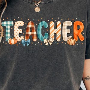 Puede incluir: Una camiseta de manga corta gris carbón con la palabra TEACHER impresa en letras grandes con varios patrones. El diseño incluye texturas de cuadros, lunares y naranja sólido, acentuado con pequeñas ilustraciones de calabazas y lazos.
