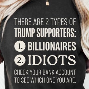 Puede incluir: Una camiseta negra con texto en blanco que dice: THERE ARE 2 TYPES OF TRUMP SUPPORTERS: 1. BILLIONAIRES 2. IDIOTS CHECK YOUR BANK ACCOUNT TO SEE WHICH ONE YOU ARE. La prenda tiene cuello redondo y mangas cortas con un diseño gráfico impreso.