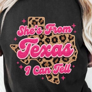 Puede incluir: Una camiseta negra con un gráfico del estado de Texas con un patrón de estampado de leopardo marrón. El texto "She's From Texas I Can Tell" aparece en una fuente redondeada de color rosa brillante. Pequeñas estrellas rosas rodean el gráfico.