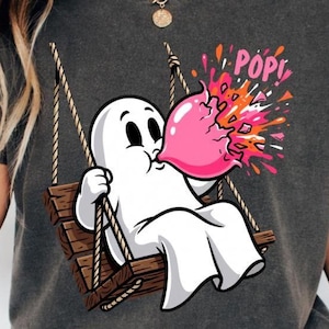 Puede incluir: Una camiseta gris carbón con un gráfico de un fantasma blanco sentado en un columpio de madera, soplando una gran burbuja de chicle rosa que estalla con la palabra POP en letras rosas, rodeada de salpicaduras coloridas tipo confeti.