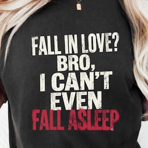 Puede incluir: Una camiseta negra de manga corta con texto blanco desgastado que dice FALL IN LOVE? BRO, I CAN'T EVEN y la palabra FALL ASLEEP impresa en letras rojas desgastadas y en negrita en el centro del área del pecho de la prenda.