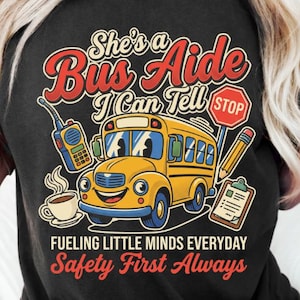 Puede incluir: Una camiseta negra con un gráfico de un autobús escolar amarillo, un walkie-talkie, una taza de café, una señal de stop, un lápiz y un portapapeles. El texto dice: She's a Bus Aide I Can Tell, Fueling Little Minds Everyday, Safety First Always.