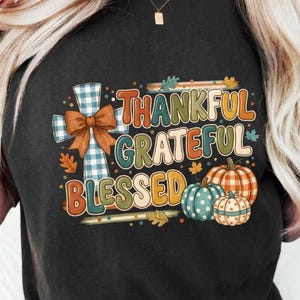 Puede incluir: Una camiseta negra de manga corta con un estampado gráfico que incluye el texto Thankful Grateful Blessed. El diseño presenta una cruz a cuadros azules y blancos con un lazo marrón y tres calabazas con patrones naranjas, azul verdoso y blancos.