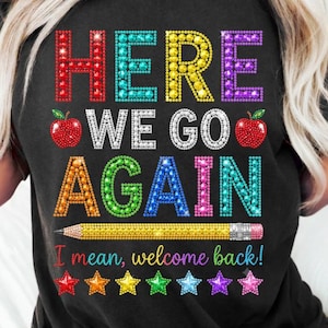 Puede incluir: Una camiseta negra con un gráfico colorido que muestra el texto "HERE WE GO AGAIN" en una fuente estilo pedrería, acompañado de dos manzanas rojas, un lápiz amarillo y una fila de seis estrellas multicolores con el texto "I mean, welcome back".